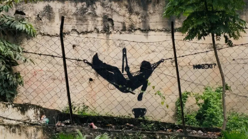 A 20-foot-long stencil graffiti art piece of the character Subodh resting on a barbed-wire hammock on a highway bridge wall in Sikkim, India, with bystanders and security personnel watching from above.